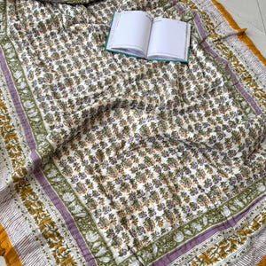 Könnte beinhalten: Eine Steppdecke mit floralem Muster mit weißem Hintergrund und grünen, lila und gelben Akzenten. Die Steppdecke hat einen Rand mit Streifenmuster und einen senfgelben Rand. Ein offenes Notizbuch liegt auf der Steppdecke.