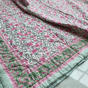 Könnte beinhalten: Eine florale Steppdecke mit weißem Hintergrund, verziert mit rosa Blüten und grünen Blättern. Die Steppdecke hat einen grün-rosa Rand mit Streifenmuster. Die Bettwäsche ist aus einem weichen, bequemen Material gefertigt.