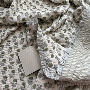 May include: A white and green floral patterned quilt with a ruffled edge. The quilt has a repeating pattern of green flowers and leaves. A notebook with lined pages is open on the quilt.