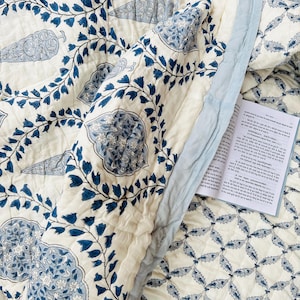 Könnte beinhalten: Eine cremefarbene Steppdecke mit blauem Blumenmuster und hellblauem Rand. Die Steppdecke weist große, stilisierte Blumen- und Blattmotive auf. Das Design umfasst ein sich wiederholendes Muster aus blauen Blättern und Blumen. Ein Buch mit Text ist sichtbar.