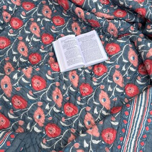 Puede incluir: Una colcha floral gris y rosa con un libro encima. La colcha tiene un patrón repetitivo de flores rosas con tallos y hojas blancas sobre un fondo gris. La colcha tiene un borde de rayas azules y blancas.