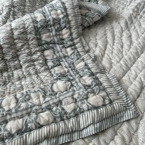 以下が含まれることがあります： グレーと白の色合いの、花柄とストライプ柄のキルトベッドスプレッド。ベッドスプレッドは、花柄モチーフとストライプのボーダーを組み合わせた詳細なデザインで、質感とレイヤー感のある外観を作り出しています。