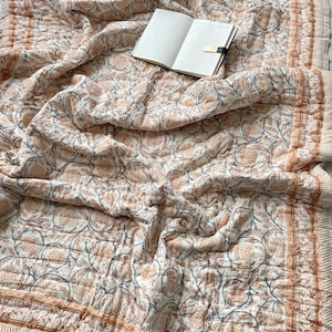 Puede incluir: Un edredón de color crema con un estampado floral en tonos melocotón y gris. El edredón tiene un borde a rayas y un cuaderno con una cubierta negra y un marcador dorado abierto encima.