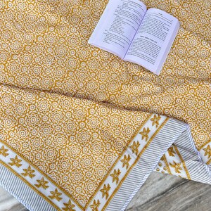 Könnte beinhalten: Eine senfgelbe Baumwolltischdecke mit einem weißen Blumenmuster und einem weißen Rand mit einem grauen Streifen und einem weißen Blumenmuster. Ein Buch liegt offen auf der Tischdecke.