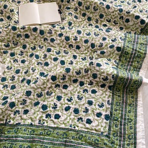 May include: A white and green floral patterned quilt with a notebook. The quilt features a repeating pattern of teal flowers and green vines on a white background. The border is a darker green with a detailed design. The notebook is open, displaying blank pages.