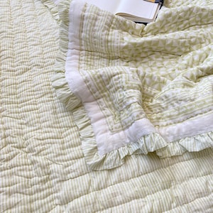 May include: A light green and white patterned quilt with ruffled edges. The quilt features a striped pattern on the main body and a geometric design on the border. An open notebook with a gold bookmark rests on the quilt.