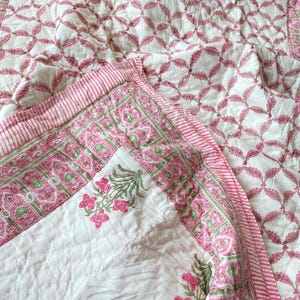 Peut inclure: Une courtepointe florale blanche et rose avec un motif de feuilles répétitif. La courtepointe a une bordure rayée rose et un motif floral détaillé sur le coin. La courtepointe est en tissu matelassé doux.