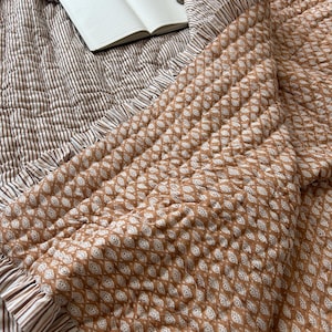 May include: A brown and white patterned quilt with a frilled edge. The quilt has a diamond pattern on one side and a striped pattern on the other. An open notebook rests on the quilt.