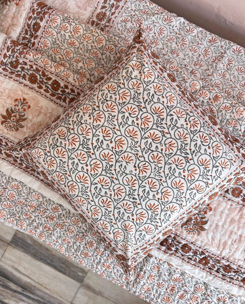 May include: A white and orange floral patterned pillow with a white background and orange and gray floral designs. The pillow is on top of a white and orange floral patterned quilt.