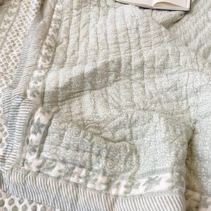 Puede incluir: Una colcha acolchada de color verde salvia y blanco con un patrón repetitivo de estampado floral y geométrico con un borde a rayas. Un cuaderno abierto con páginas blancas descansa sobre la esquina doblada de la tela acolchada.
