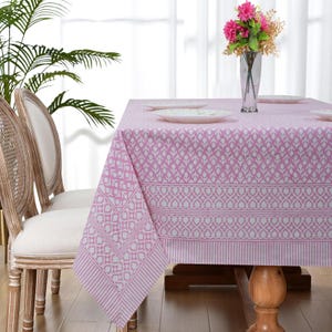 May include: A pink and white patterned tablecloth covers a wooden table, set with white plates and a vase of pink flowers. The tablecloth features a repeating floral and heart design, with a striped border. Two chairs are visible on the left.