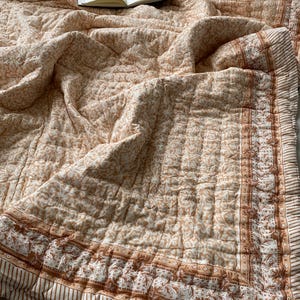 May include: A peach and cream coloured quilted bedspread with a repeating floral pattern. The border has a brown and white striped design. An open book rests on the bedspread.