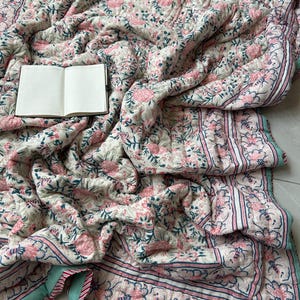 Puede incluir: Una manta acolchada con un patrón floral repetitivo en tonos rosa, verde y azul oscuro sobre un fondo crema, con un borde a rayas y un pequeño cuaderno abierto con páginas blancas en blanco descansando sobre los pliegues de la tela.