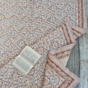 May include: A quilted textile featuring a repeating floral pattern in muted orange and grey tones on a cream background, finished with a striped border. An open book with printed text rests on the surface of the folded fabric.