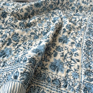 May include: A white and blue floral patterned quilt with a book on top. The quilt features a repeating floral design in shades of blue against a white background. The edges have a striped border.