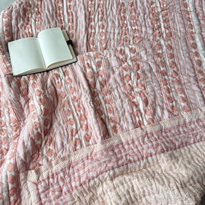 Può includere: Una coperta trapuntata con un motivo geometrico rosa e bianco. La coperta presenta strisce verticali e forme a diamante. Un taccuino aperto con pagine bianche poggia sulla coperta.
