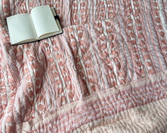 May include: A quilted blanket with a pink and white geometric pattern. The blanket features vertical stripes and diamond shapes. An open notebook with blank pages rests on the blanket.