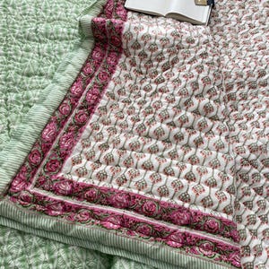 Könnte beinhalten: Eine gesteppte Decke mit weißem Grund und wiederkehrendem grün-rosa Blumenmuster, umrandet von einem breiten Band aus rosa Rosen und grünen Blättern, mit einem hellgrünen Blattmuster-Abschnitt und einem offenen Notizbuch darauf.