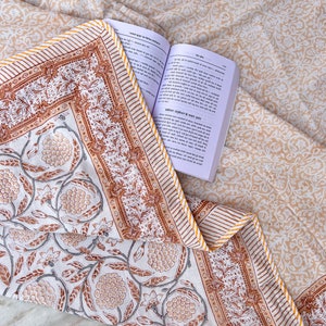 May include: A white cotton quilt with a brown and yellow floral print. The quilt has a yellow and white striped border. A book with text in a foreign language is lying on top of the quilt.