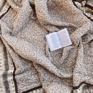 Può includere: Un trapunta con motivo marrone e bianco con un bordo marrone. La trapunta è piegata e ha un libro sopra.