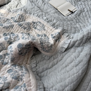 Puede incluir: Un edredón con estampado floral azul claro y blanco con bordes con volantes. El edredón está sobre una cama con una colcha estampada a juego. Un cuaderno abierto con un clip dorado descansa sobre la colcha.
