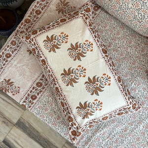 May include: A white pillowcase with a brown floral pattern. The pillowcase has a border of brown and orange flowers. The pillowcase is on top of a white bedspread with a brown floral pattern.
