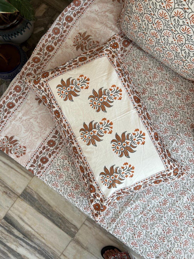 May include: A white pillowcase with a brown floral pattern. The pillowcase has a border of brown and orange flowers. The pillowcase is on top of a white bedspread with a brown floral pattern.