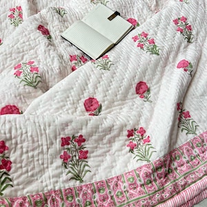 Könnte beinhalten: Weiß gesteppte Decke mit einem Blumenmuster in Rosa und Grün. Die Decke hat ein sich wiederholendes Design aus stilisierten Blumen und Blättern. Ein schwarzes Notizbuch mit goldenem Verschluss liegt offen auf der Decke.