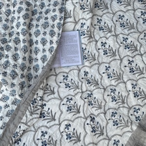 May include: A white and blue floral patterned quilt with a book open on top. The quilt features a repeating pattern of blue flowers and gray leaves on a white background. The quilt is folded over, revealing a different floral pattern.