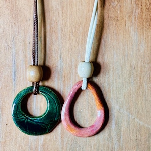 Peut inclure: Deux pendentifs en céramique sur cordons. L'un est un pendentif circulaire émaillé vert foncé sur un cordon marron avec une perle en bois. L'autre est un pendentif irrégulier émaillé rose et orange sur un cordon blanc avec une perle en bois clair.
