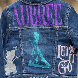 May include: A blue denim jacket with a purple glitter design that says "AUBREE" on the back. The front of the jacket has a silver glitter Olaf design, a turquoise glitter Elsa design, and pink glitter text that says "Let it Go" and "FROZEN".