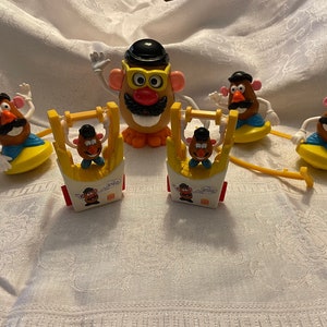 May include: A set of Mr. Potato Head toys, including a large Mr. Potato Head with glasses and a mustache, and five smaller Mr. Potato Head figures. The smaller figures are riding on various vehicles, including a yellow boat, a yellow car, and a yellow swing set.