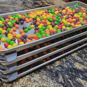 May include: A stack of four metal trays filled with colorful candies. The candies are in various colors, including red, orange, yellow, green, and purple. The trays are stacked on top of each other, and the candies are evenly distributed across the surface of each tray.