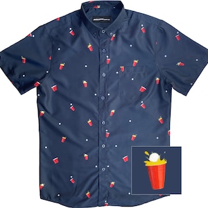 May include: A navy blue short-sleeved button-down shirt with a red solo cup pattern. The shirt has a pocket on the left side and a white button-up closure. The shirt has a small image of a red solo cup with a golf ball in it.