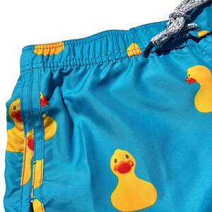 May include: Blue swim trunks with a pattern of yellow rubber duckies. The trunks have a drawstring waist and a side seam.