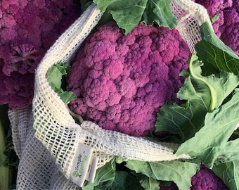 Puede incluir: Coliflor morada en una bolsa de malla blanca con fondo de hojas verdes. La bolsa tiene una etiqueta marrón que dice "Simple Ecology".