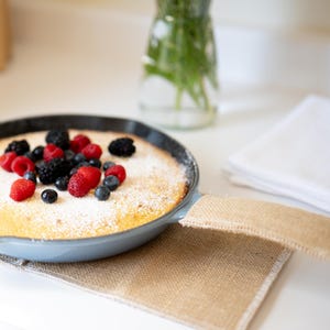 May include: A light blue cast iron skillet containing a golden-brown pancake topped with fresh raspberries, blackberries, and blueberries, dusted with white powdered sugar, resting on a tan burlap fabric handle cover.