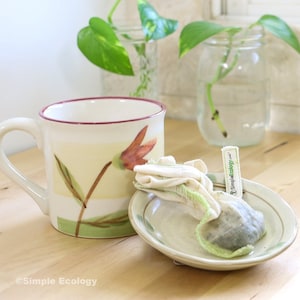 May include: A white ceramic mug with a floral design, a small saucer, and a reusable tea bag in a white cloth pouch with a green label that says "SimpleEcology.com".
