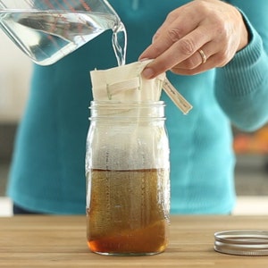 May include: A person pouring water into a glass jar with a white cloth filter inside. The jar is filled with a dark brown liquid.