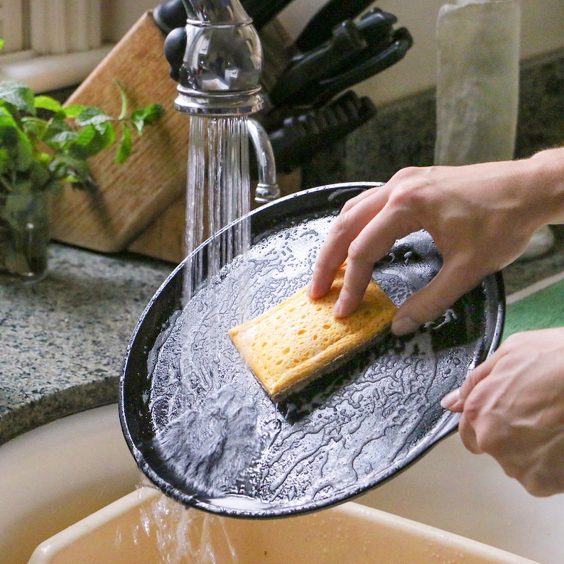May include: A round black cast iron skillet being cleaned under a kitchen faucet with a yellow rectangular sponge. The skillet is held at an angle while water flows over the surface, creating soapy bubbles and rinsing the dark metal cookware.