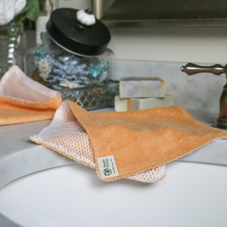 May include: Two orange and white dishcloths with a mesh backing. The dishcloths have a green label that says "Simple Ecology" and a website address. The dishcloths are sitting on a white countertop next to a sink.