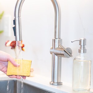 May include: A stainless steel high-arc kitchen faucet with a pull-down sprayer head dispensing water onto a rectangular yellow sponge. Next to the faucet sits a clear glass soap dispenser with a stainless steel pump mechanism on a white counter.