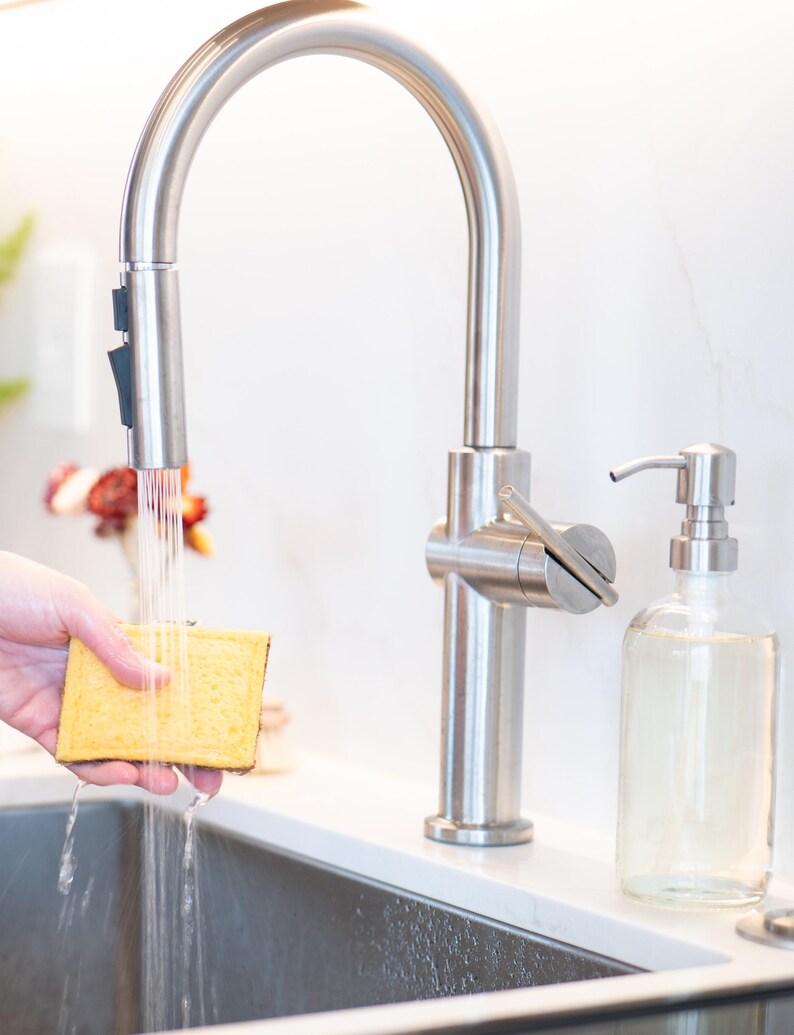 May include: A stainless steel high-arc kitchen faucet with a pull-down sprayer head dispensing water onto a rectangular yellow sponge. Next to the faucet sits a clear glass soap dispenser with a stainless steel pump mechanism on a white counter.
