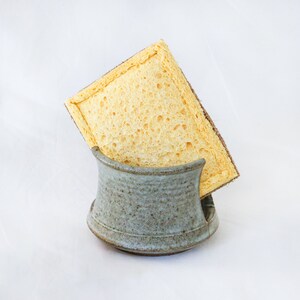 May include: A rectangular yellow cellulose sponge with a brown fibrous scouring layer on one side, resting inside a small, speckled, light blue-grey ceramic holder with a curved, open-front design and a wide, stable base for countertop storage.