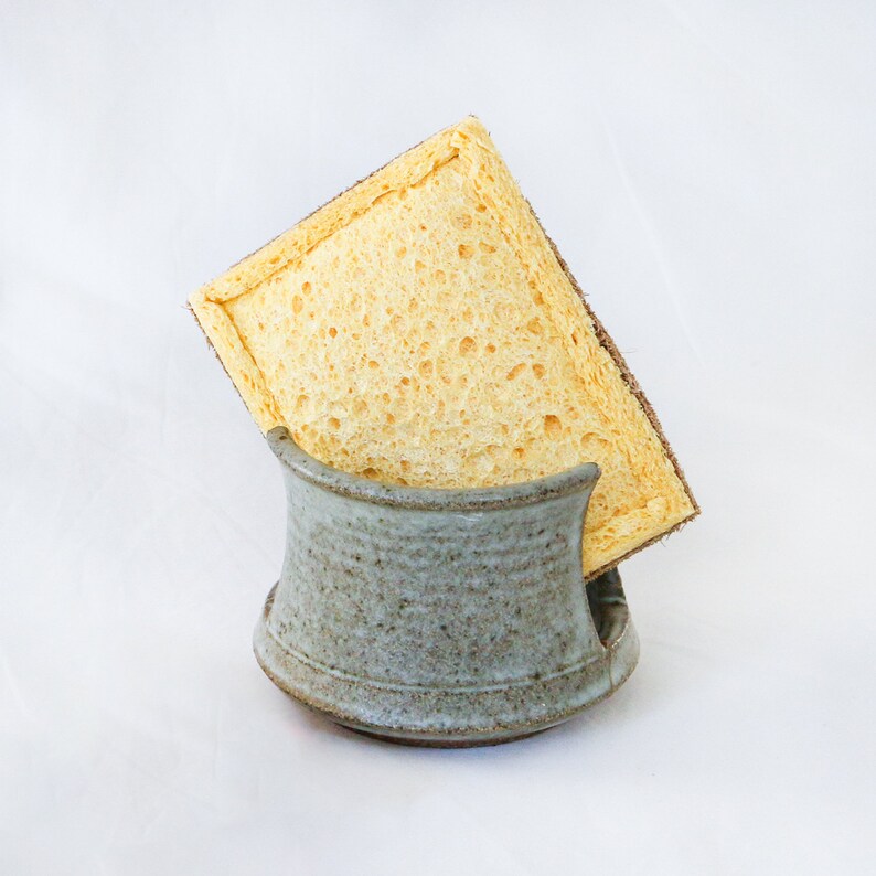 May include: A rectangular yellow cellulose sponge with a brown fibrous scouring layer on one side, resting inside a small, speckled, light blue-grey ceramic holder with a curved, open-front design and a wide, stable base for countertop storage.