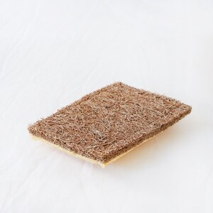 May include: A rectangular scouring pad featuring a thick, brown, fibrous top layer with a coarse, tangled texture and a thinner, light beige, dense sponge base layer. The object is designed for scrubbing surfaces and cleaning various household items.