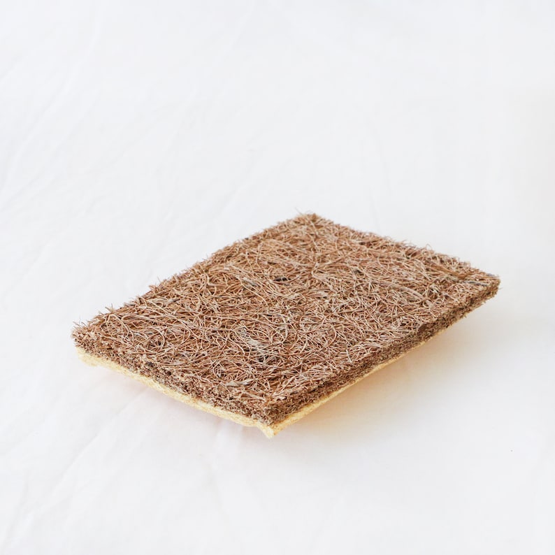 May include: A rectangular scouring pad featuring a thick, brown, fibrous top layer with a coarse, tangled texture and a thinner, light beige, dense sponge base layer. The object is designed for scrubbing surfaces and cleaning various household items.