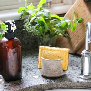 May include: A ceramic sponge holder with a speckled beige finish holding a rectangular yellow sponge. The holder has a curved front design and is positioned next to a dark amber glass soap dispenser with a black pump and a chrome faucet.