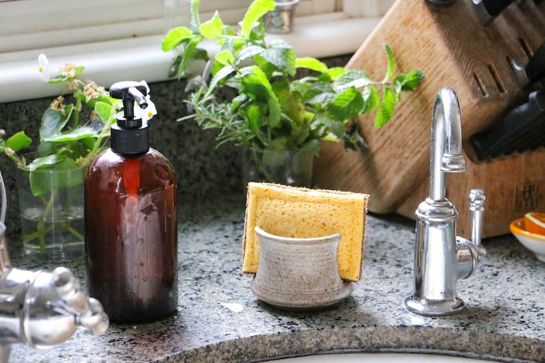 May include: A ceramic sponge holder with a speckled beige finish holding a rectangular yellow sponge. The holder has a curved front design and is positioned next to a dark amber glass soap dispenser with a black pump and a chrome faucet.
