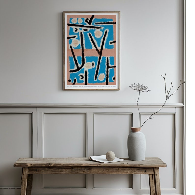 May include: A framed abstract art print with black lines and shapes on a blue and pink background. The print is hanging on a white wall with a wooden table and a vase with dried flowers in front of it.